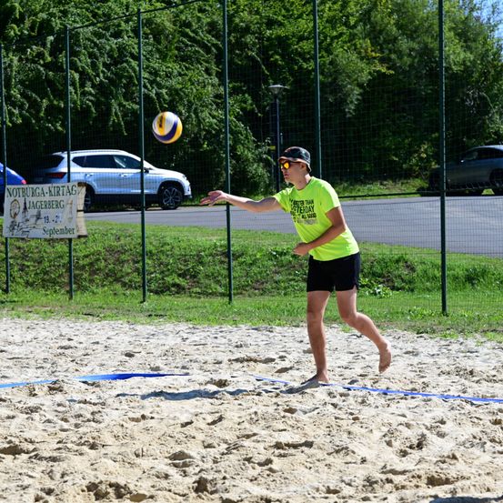 Eine Person spielt Volleyball auf einem sandigen Platz, trägt ein gelbes T-Shirt und schwarze Shorts. Ein Netz und ein Banner sind hinter ihm.