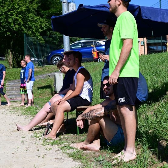Eine Gruppe von Männern sitzt unter einem Regenschirm, einer steht in der Nähe. Sie tragen Freizeitkleidung und scheinen sich in einem Park zu befinden. Hinter ihnen ist ein Auto geparkt und eine Hecke im Hintergrund.