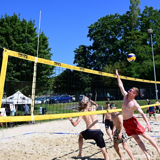 Eine Gruppe von Männern spielt Beachvolleyball auf einem Sandplatz. Ein Mann springt, um den Ball zu schlagen, während ein anderer zuschaut. Dahinter steht ein gelbes Netz mit schwarzem Mesh und einigen Bäumen.