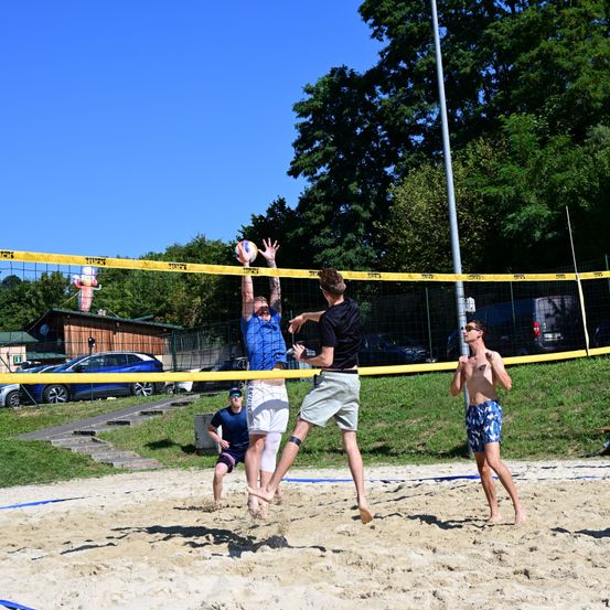 Eine Gruppe von Männern spielt Volleyball auf einem sandigen Platz mit gelben Netzen. Ein Mann springt, um den Ball zu schlagen, während andere sich darauf vorbereiten, zu blocken.