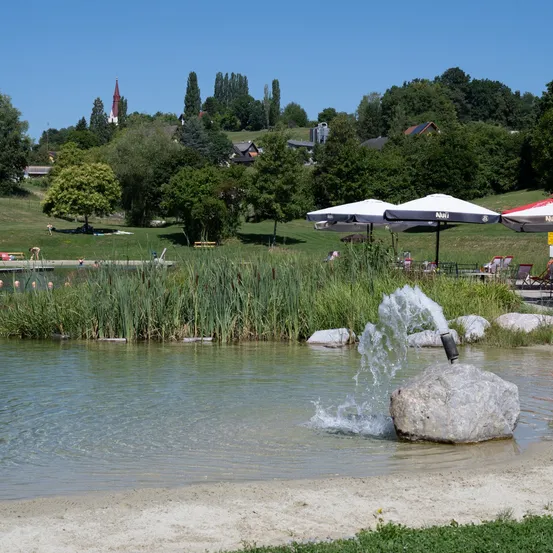 Ein ruhiger See mit einem Brunnen, umgeben von Steinen und Gras, mit Menschen, die sich unter Sonnenschirmen am Ufer entspannen, und einer Kulisse aus Bäumen und einem entfernten Kirchturm.