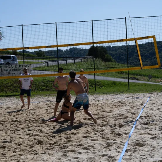 Vier Männer spielen Beachvolleyball auf einem sandigen Spielfeld, wobei eine Frau gefallen ist. Ein Zaun umgibt das Spielfeld und ein Schild befindet sich am Zaun.