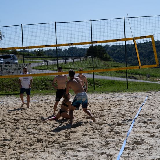 Vier Männer spielen Beachvolleyball auf einem sandigen Spielfeld, wobei eine Frau gefallen ist. Ein Zaun umgibt das Spielfeld und ein Schild befindet sich am Zaun.