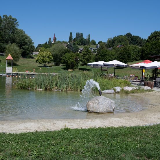 Ein ruhiger See mit einem Springbrunnen, einem großen Felsen und einigen schwimmenden Personen. Bäume und ein Turm in der Ferne bilden den Hintergrund.