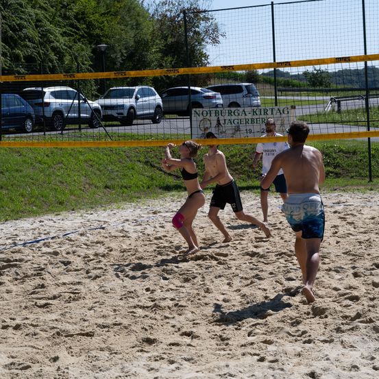Eine Gruppe von Menschen spielt Volleyball auf einem Sandplatz. Ein Netz trennt die beiden Teams. Ein Banner mit Text hängt über dem Spielfeld.