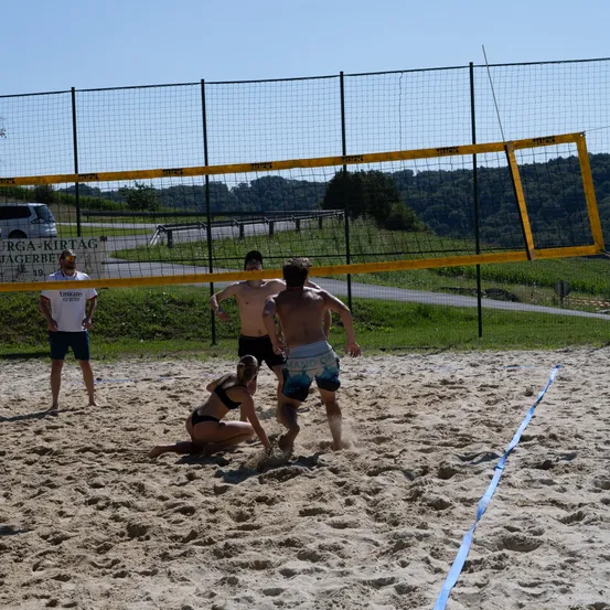 Eine Gruppe von Menschen spielt Volleyball auf einem sandigen Platz. Zwei Männer springen, um den Ball zu schlagen, und eine Frau hockt sich, um ihn zu empfangen. Dahinter steht ein gelbes Netz und ein Zaun. Ein Mann in einem weißen Shirt steht in der Nähe des Zauns.