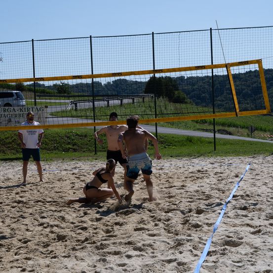 Eine Gruppe von Menschen spielt Volleyball auf einem sandigen Platz. Zwei Männer springen, um den Ball zu schlagen, und eine Frau hockt sich, um ihn zu empfangen. Dahinter steht ein gelbes Netz und ein Zaun. Ein Mann in einem weißen Shirt steht in der Nähe des Zauns.