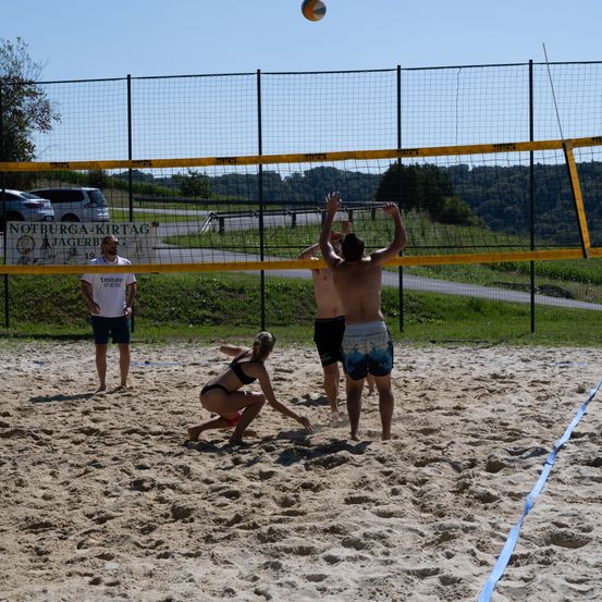 Eine Gruppe von Menschen spielt Volleyball auf einem sandigen Platz. Zwei Männer springen und greifen nach dem Ball, während eine Frau hockt, um zu blocken. Ein Zaun umgibt das Feld.