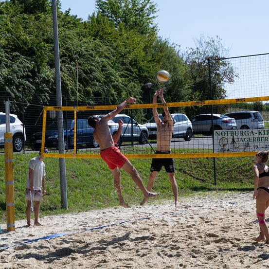 Zwei Männer spielen Volleyball auf einem Sandplatz, einer springt, um den Ball zu schlagen, während der andere blockt. Zuschauer beobachten in der Nähe.