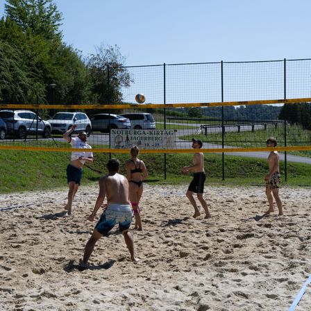 Sechs Personen spielen Beachvolleyball in einem eingezäunten Bereich. Ein Banner mit den Worten 'Notburga-Kirttag Jagerberg' ist am Zaun angebracht. Autos sind im Parkplatz geparkt.