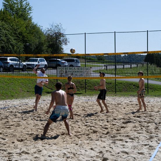 Sechs Personen spielen Beachvolleyball in einem eingezäunten Bereich. Ein Banner mit den Worten 'Notburga-Kirttag Jagerberg' ist am Zaun angebracht. Autos sind im Parkplatz geparkt.