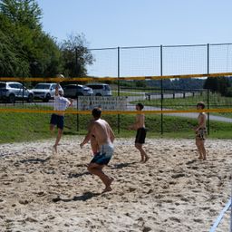 Fünf Männer spielen Beachvolleyball auf einem sandigen Platz. Ein Spieler springt, um den Ball zu schlagen. Ein weiterer Spieler beobachtet hinter dem Netz. Der Platz ist von einem Metallzaun umschlossen.