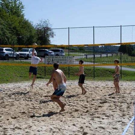 Fünf Männer spielen Beachvolleyball auf einem sandigen Platz. Ein Spieler springt, um den Ball zu schlagen. Ein weiterer Spieler beobachtet hinter dem Netz. Der Platz ist von einem Metallzaun umschlossen.