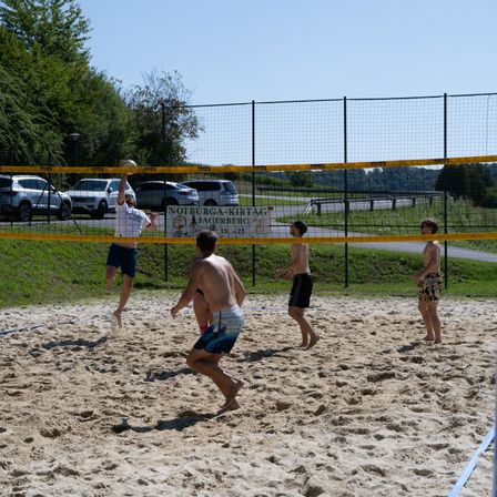 Fünf Männer spielen Beachvolleyball auf einem sandigen Platz. Ein Spieler springt, um den Ball zu schlagen. Ein weiterer Spieler beobachtet hinter dem Netz. Der Platz ist von einem Metallzaun umschlossen.