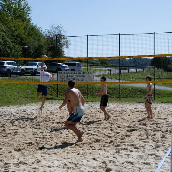 Fünf Männer spielen Beachvolleyball auf einem sandigen Platz. Ein Spieler springt, um den Ball zu schlagen. Ein weiterer Spieler beobachtet hinter dem Netz. Der Platz ist von einem Metallzaun umschlossen.