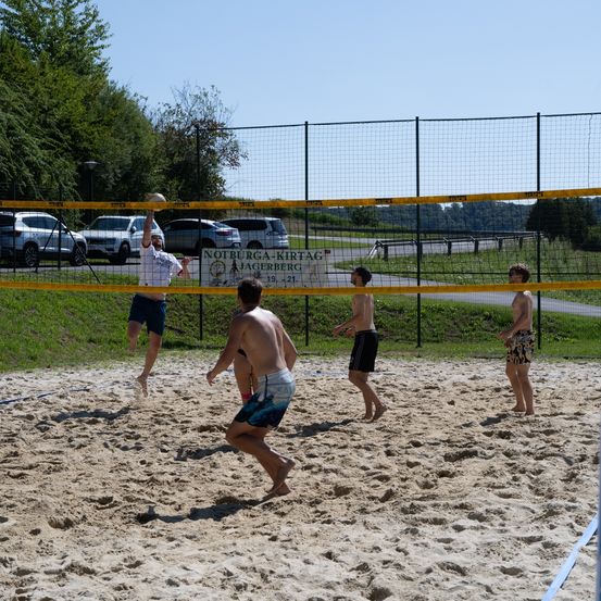 Fünf Männer spielen Beachvolleyball auf einem sandigen Platz. Ein Spieler springt, um den Ball zu schlagen. Ein weiterer Spieler beobachtet hinter dem Netz. Der Platz ist von einem Metallzaun umschlossen.