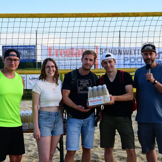 Fünf Personen stehen auf einem sandigen Strand, wahrscheinlich bei einem Volleyball-Event. Ein Mann hält eine Schachtel Makava Eistee. Ein Volleyballnetz und Werbebanner sind im Hintergrund zu sehen.