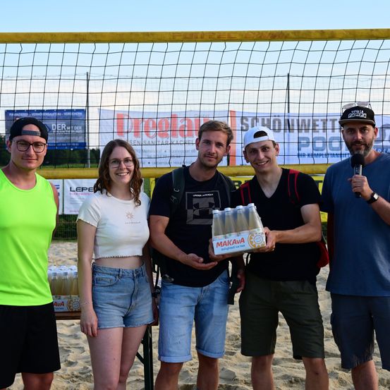 Fünf Personen stehen auf einem sandigen Strand, wahrscheinlich bei einem Volleyball-Event. Ein Mann hält eine Schachtel Makava Eistee. Ein Volleyballnetz und Werbebanner sind im Hintergrund zu sehen.