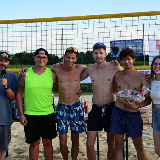 Fünf Männer und eine Frau stehen auf einem Strand in der Nähe eines Volleyballnetzes. Ein Mann hält ein Mikrofon, ein anderer ein verpacktes Geschenk. Sie alle lächeln in die Kamera.