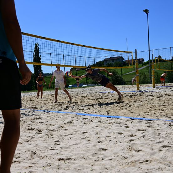 Eine Gruppe von Menschen spielt an einem sonnigen Tag Beachvolleyball. Ein Mann taucht, um den Ball zu schlagen.