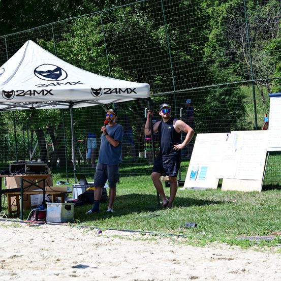 Zwei Männer stehen unter einem Zelt mit 24MX-Marke, einer hält ein Mikrofon und der andere trägt eine Schutzbrille. Ein Zaun, Bäume und eine Person sind im Hintergrund zu sehen.