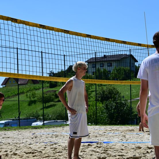 Eine Gruppe von Menschen spielt Volleyball auf einem sandigen Platz mit einem Netz. Eine Person steht vor dem Netz und schaut sich den Ball an.