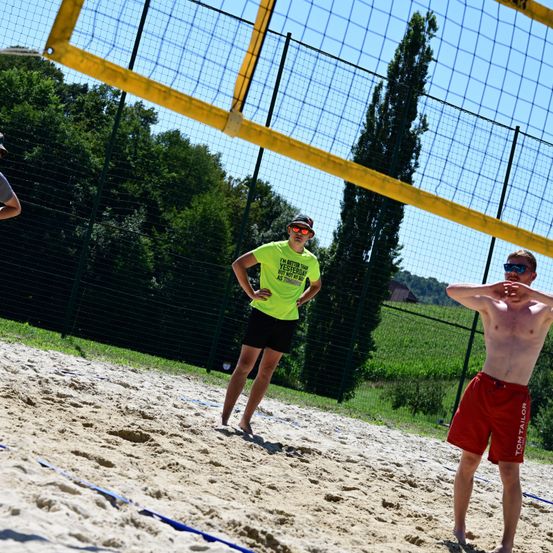 Zwei Personen spielen Beachvolleyball auf einem sandigen Platz, mit einem Netz in der Mitte. Eine Person trägt ein gelbes Shirt und eine Sonnenbrille, während die andere Oberkörper frei hat und rote Shorts trägt. Bäume und ein Zaun sind im Hintergrund zu sehen.