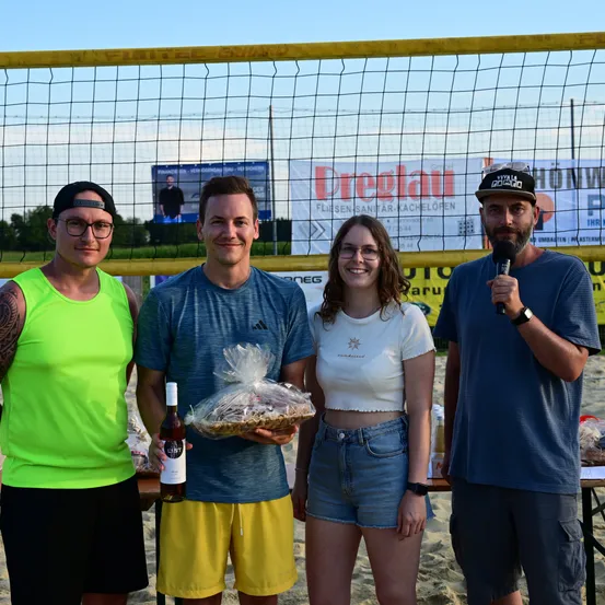 Vier Personen stehen lächelnd vor einem Beachvolleyballnetz. Ein Mann hält ein Mikrofon und eine Geschenktüte, ein anderer eine Flasche. Die Frau in der Mitte trägt ein weißes T-Shirt und Jeansshorts. Dahinter befindet sich ein Banner mit Text.