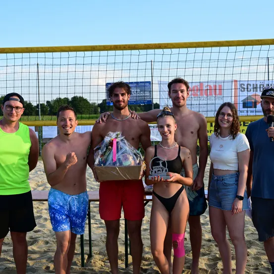 Eine Gruppe von Menschen steht auf einem sandigen Strand und posiert mit einem Pokal und einer Geschenkbox. Ein Volleyballnetz ist im Hintergrund. Sie lächeln und scheinen einen Sieg zu feiern.