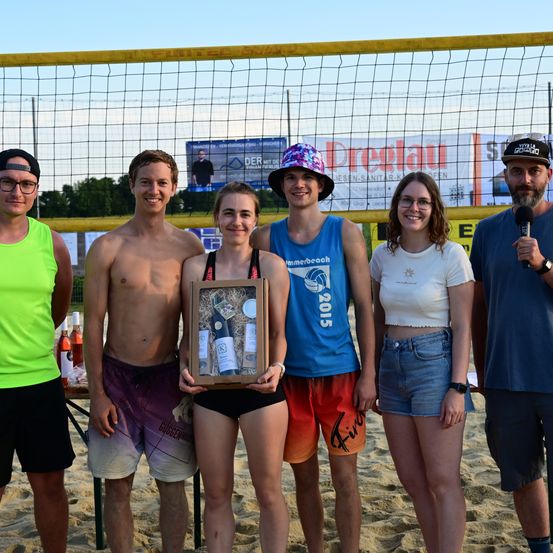 Eine Gruppe von fünf Personen steht am Strand, wobei eine Person eine Geschenkbox hält. Die Frau in der Mitte trägt einen Badeanzug und hält einen gerahmten Preis. Hinter ihnen ist ein Beachvolleyballnetz zu sehen.