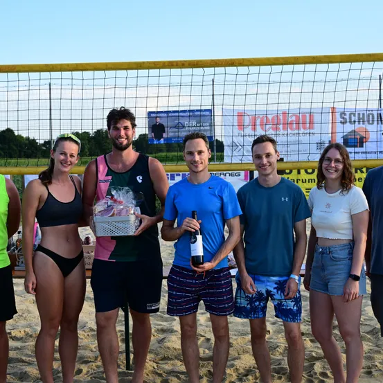 Eine Gruppe von sechs Personen, darunter drei Männer und drei Frauen, steht auf einem sandigen Strand mit einem Volleyballnetz im Hintergrund. Sie halten Geschenke und eine Flasche. Sie alle lächeln für die Kamera.