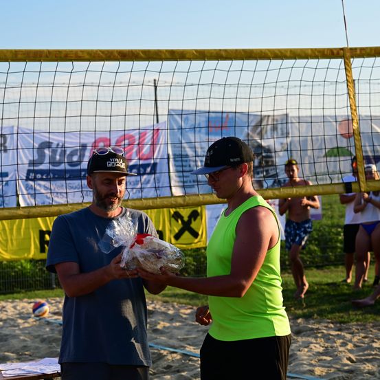 Ein Mann in einem blauen T-Shirt überreicht einem Mann in einem grünen Tanktop einen eingewickelten Gegenstand vor einem Beachvolleyball-Netz.