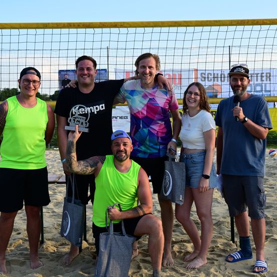 Eine Gruppe von Menschen steht auf einem Strand vor einem Volleyballnetz, lächelt und posiert für ein Foto. Ein Mann hält eine Trophäe und ein Mikrofon. Sie tragen lässige Sommerkleidung.