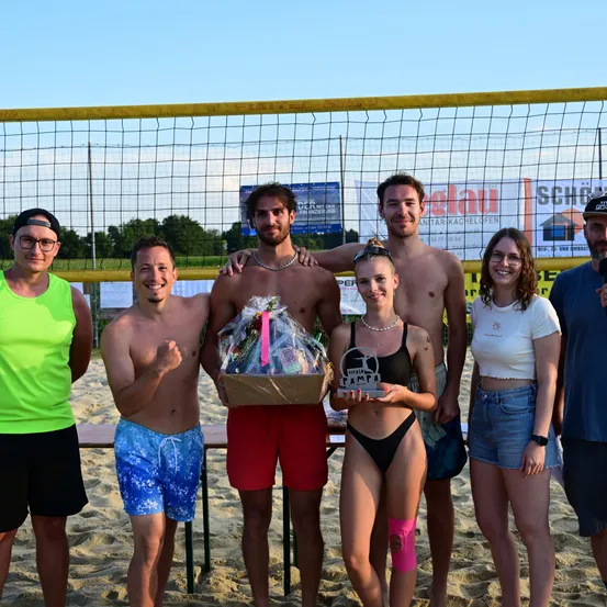 Eine Gruppe junger Erwachsener posiert für ein Foto am Strand mit einem Volleyballnetz im Hintergrund. Eine Frau hält einen Pokal und einen Geschenkkorb, während andere lässige Kleidung tragen.