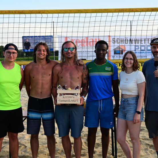Fünf Personen stehen auf einem Strand mit einem Netz im Hintergrund und lächeln. Ein Mann hält eine Box mit Schokoladen. Ein anderer Mann spricht in ein Mikrofon.