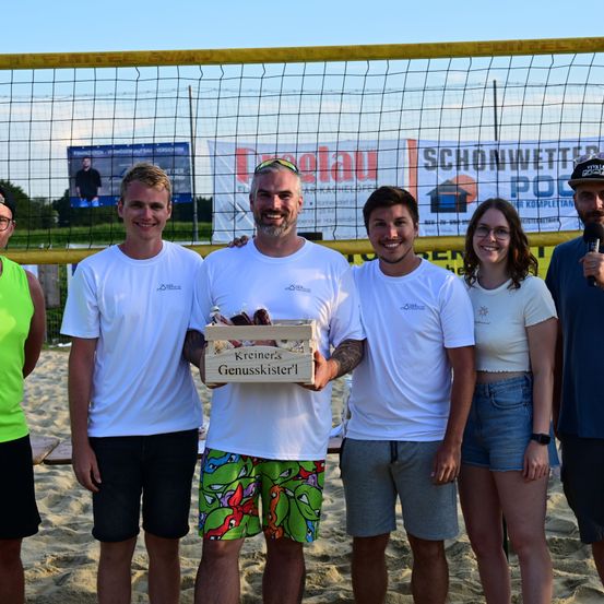 Fünf Personen stehen lächelnd auf einem Strand. Der Mann in der Mitte hält eine Box mit der Aufschrift 'Kreiners Genussskiller'. Hinter ihnen steht ein Volleyballnetz und eine Werbetafel.