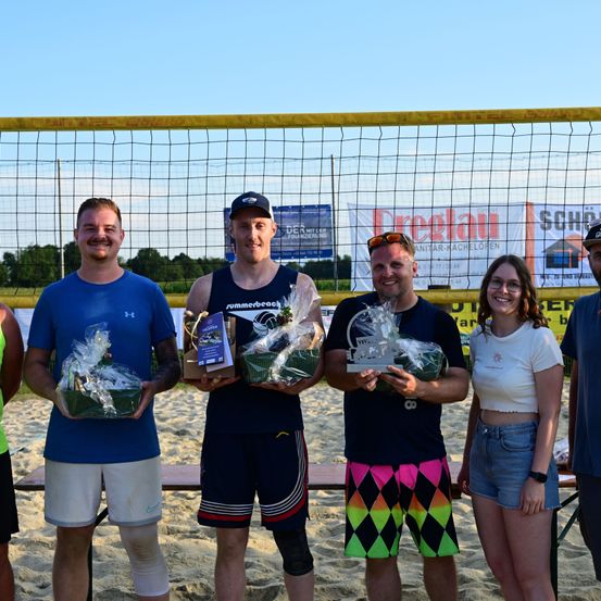 Eine Gruppe von Personen steht auf einer sandigen Oberfläche und hält eingewickelte Geschenke. Sie lächeln und scheinen zu feiern. Ein Volleyballnetz und Banner befinden sich hinter ihnen.