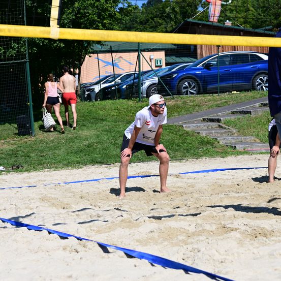 Ein Mann in einem weißen T-Shirt und schwarzen Shorts spielt Volleyball auf einem sandigen Spielfeld mit einem Netz, während andere auf einem Grasfeld hinter ihm spazieren.