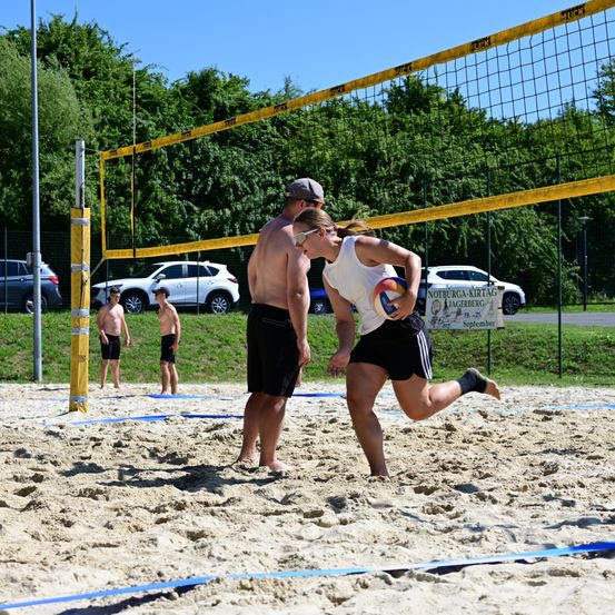 Zwei Personen spielen Volleyball auf einem Sandplatz mit Netz. Eine Person läuft und hält den Ball, während eine andere Person hinter dem Netz steht. Ein Banner und Autos sind im Hintergrund.