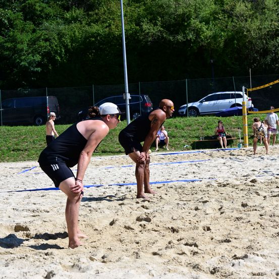 Zwei Männer spielen Beachvolleyball auf einem sandigen Platz mit Netz, während mehrere Personen auf dem Rasen sitzen. Autos sind hinter einem Zaun geparkt.