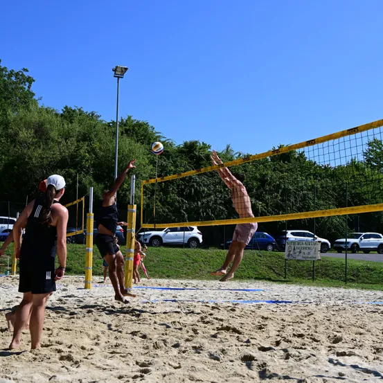 Eine Gruppe von Menschen spielt Volleyball auf einem sandigen Strand. Eine Person springt, um den Ball über das Netz zu schlagen, während andere zusehen.