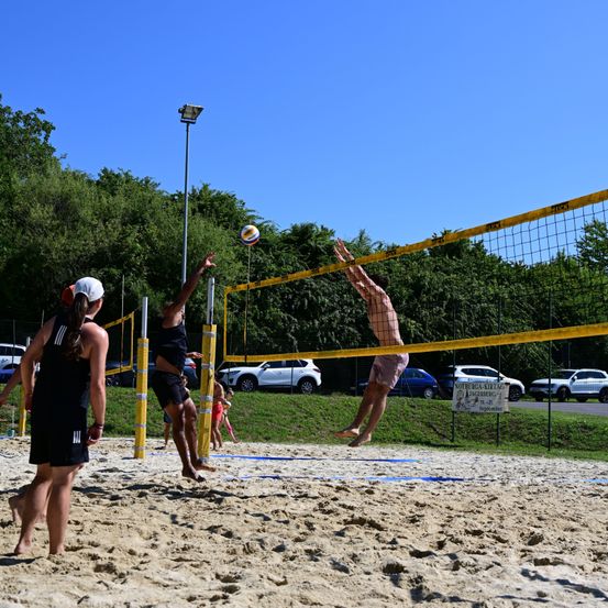 Eine Gruppe von Menschen spielt Volleyball auf einem sandigen Strand. Eine Person springt, um den Ball über das Netz zu schlagen, während andere zusehen.