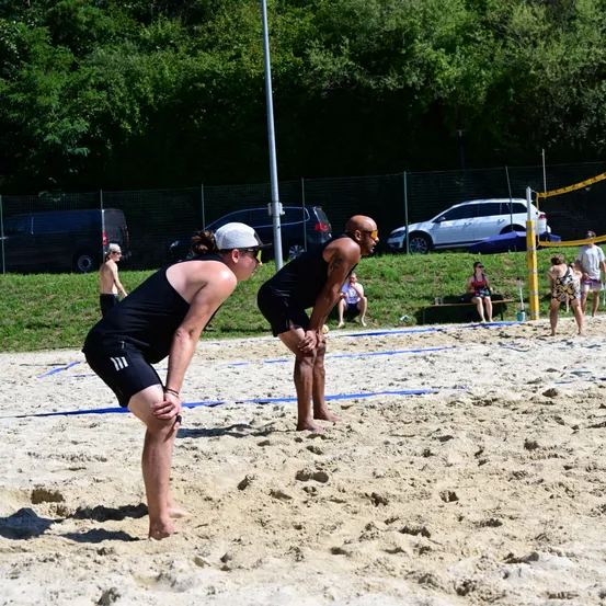Zwei Männer in schwarzen Tanktops und Shorts spielen Beachvolleyball auf einem sandigen Spielfeld, wobei sie durch ein Netz getrennt sind. Zuschauer sitzen und stehen um den Rand herum. Dahinter sind Autos in der Nähe eines Zauns geparkt, und es gibt Bäume und Gras im Hintergrund.