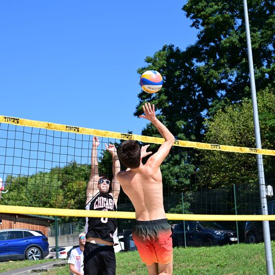Zwei junge Männer spielen Volleyball an einem sonnigen Tag. Einer springt, um zu blocken, während der andere sich darauf vorbereitet, zu schlagen. Das Spielfeld ist von einem Rasenfeld und Bäumen umgeben.