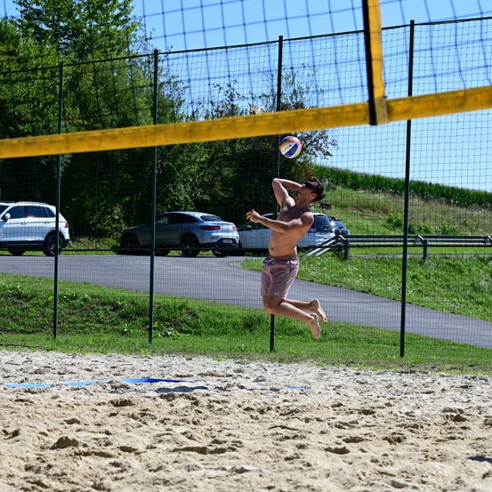 Ein oberkörperfreier Mann springt über ein Netz, um einen Volleyball auf einem Sandstrand zu schlagen, mit einem Weg und geparkten Autos im Hintergrund.