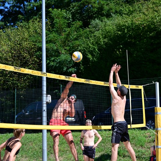 Zwei Männer spielen Volleyball an einem sonnigen Tag, einer in roten Shorts und der andere ohne Hemd. Eine Frau in einem schwarzen Bikini und ein Junge beobachten in der Nähe. Bäume und ein Auto sind im Hintergrund.