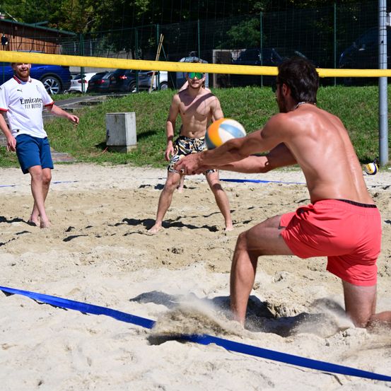 Drei Männer spielen Beachvolleyball auf einem Sandplatz. Der Mann in roten Shorts springt, um den Ball zu schlagen. Ein anderer Mann in einer Sonnenbrille beobachtet.