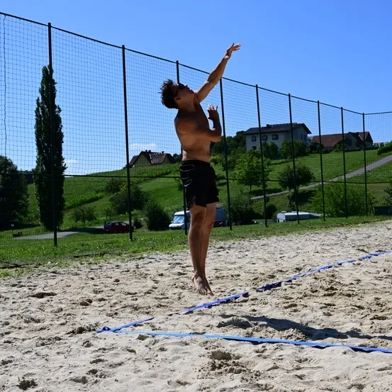 Ein Mann in schwarzen Shorts springt und streckt seine Hände in Richtung Volleyballnetz. Das Beach-Court ist von einem Zaun umgeben. Bäume und Häuser sind im Hintergrund sichtbar.