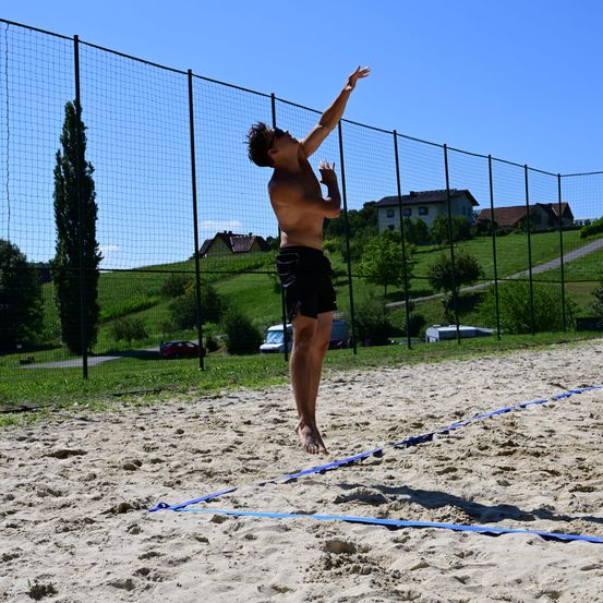 Ein Mann in schwarzen Shorts springt und streckt seine Hände in Richtung Volleyballnetz. Das Beach-Court ist von einem Zaun umgeben. Bäume und Häuser sind im Hintergrund sichtbar.