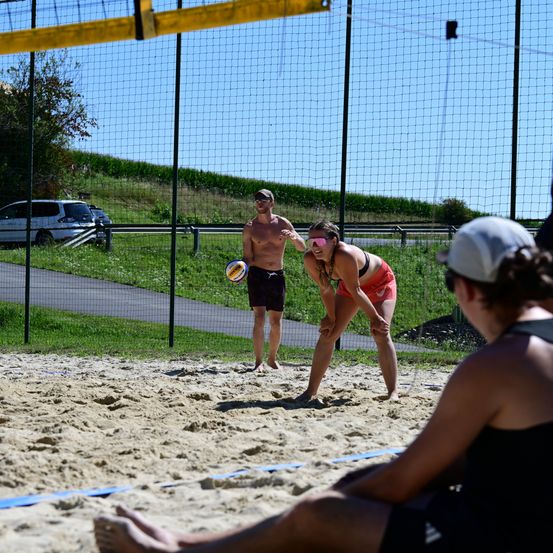Drei Personen spielen Volleyball auf einem Sandplatz. Ein Mann steht bereit zum Aufschlag, während eine Frau den Ball schlagen will. Eine weitere Person sitzt im Sand und beobachtet das Spiel.